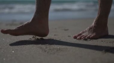 Yürüyen, yürüyen ve deniz sahilindeki altın kumda duran bir adam. Süper yavaş çekim. Okyanus kıyısında yürürken erkek bacakları alçak açıyla çekiliyor. Yalınayak dalgalarla kumlu sahilde. Yaz kıyı şeridi.