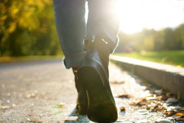 Kız sonbahar şehir parkında yol boyunca yürür. Bir kızın bacaklarında sonbahar kadın ayakkabıları. Kız kot pantolon giyiyor..