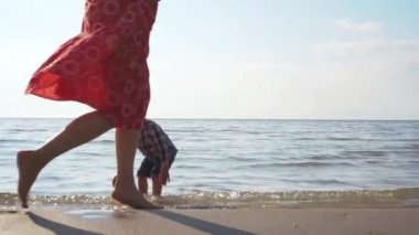 Yavaş çekim bir kadının kumsalda yürüdüğünü gösteriyor. Oğlunun su ve kumda oynadığını. Sahilde oynayan mutlu ailenin destansı gösterisi. Mutlu aile birlikteliği kavramı tatilde dışarıda..