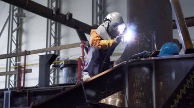 Koruyucu maskeli ve takım elbiseli profesyonel ağır endüstri işçisi fabrikada metal yapımında çalışıyor. Çelik ve demir kaynaklama şantiyesi. Kaynak makinesi kıvılcımlar ve parıltılarla.