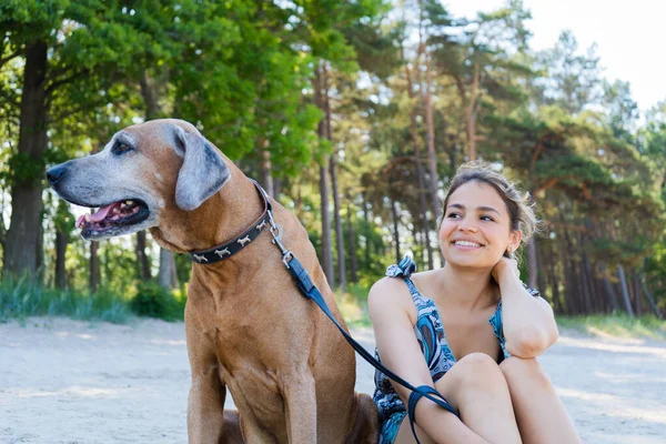 Güzel genç bir kız köpeğiyle sahilde yalnız oturuyor. Şirin, gülümseyen kadın ve Rodezyalı sırt hayvanı en iyi arkadaşlardır..