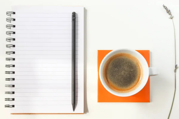 白いテーブルの上にオレンジのコースターの上にコーヒーのカップ その隣にノートとラベンダーの枝 作業机 — ストック写真