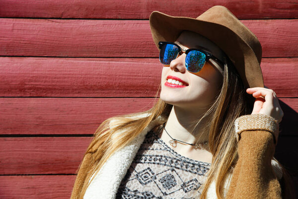 Young beautiful caucasian smiling girl in brown hat, sunglasses and kint on background wooden wall. Country style, copy space.