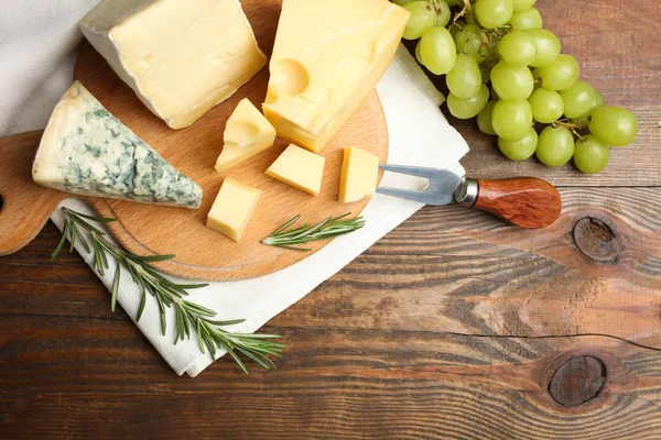 Cheese platter: yellow Maasdam cheese, white Camembert cheese and blue cheese Dor Blue with rosemary, grapes and fork on cutting board on wooden background. Top view. Concept serving cheese.
