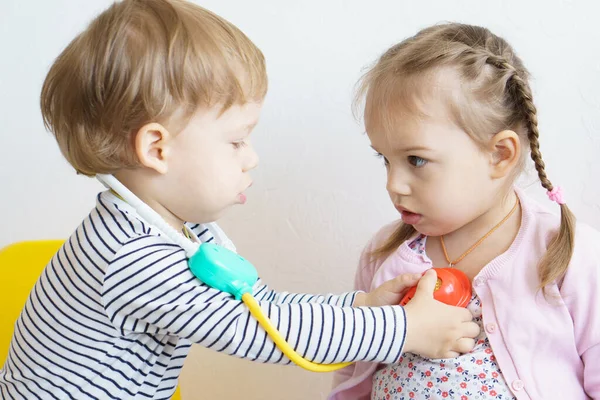 Two children play doctor in the nursery. Brother in yellow glasses treats his sister, playing dentist, ENT, ophthalmologist, temperature measurement. Role-playing games for children, the development Stock Image
