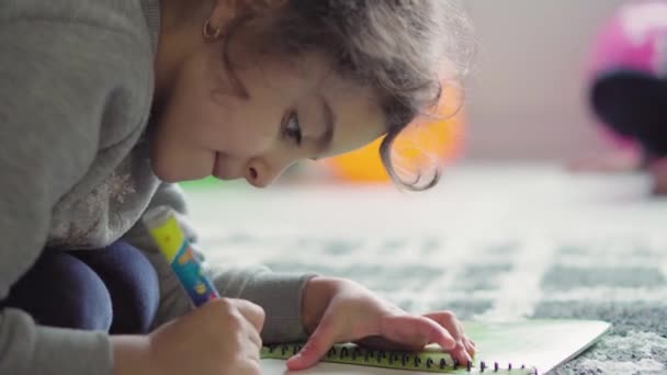 子供時代,遊び,トレーニング,自己分離概念-閉じて暗い髪巻き小さな6歳のペルシア語中東の女の子外観は、床の上に彼女の胃の上に横たわるノートブック鉛筆で描かれています. — ストック動画