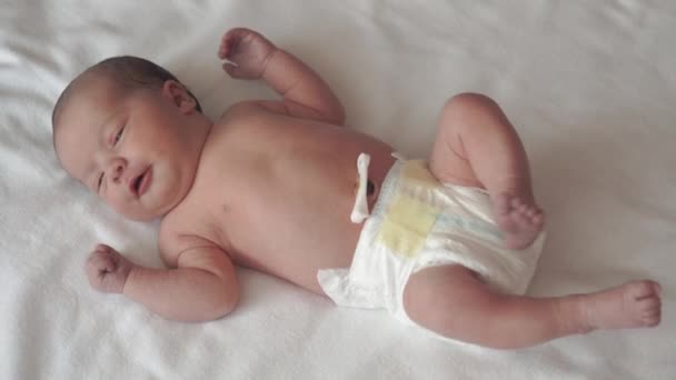 Infancy, childhood, development, medicine and health concept - close-up face of newborn naked calm awake baby grimaces lying on back with belly button holding medical clip on white beige background — Stock Video