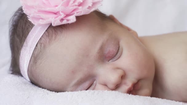 Βρεφική ηλικία, παιδική ηλικία, ανάπτυξη, ιατρική και υγειονομική έννοια - γκρο πλαν πρόσωπο ενός νεογέννητου γυμνού κοριτσιού που κοιμάται ξαπλωμένη στο στομάχι της με έναν επίδεσμο και ένα λουλούδι στο κεφάλι της σε ροζ φόντο. — Αρχείο Βίντεο