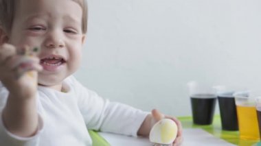 Sarışın bir çocuk tavuk yumurtalarını gıda boyasıyla boyuyor. Paskalya hazırlığı. Kapalı mekanda ellerle resim yapmak. 3 yaşından küçük çocuklar için yaratıcı bir gelişme. Eğitim, evde çizim yaparken