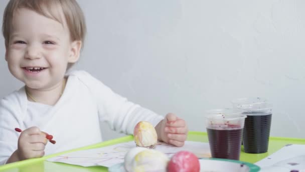 Один маленький усміхнений хлопчик малює курячі яйця харчовими барвниками. Підготовка до Великодня. Живопис руками в приміщенні. Творчий розвиток для дітей до 3 років. Освіта, малювання вдома під час — стокове відео