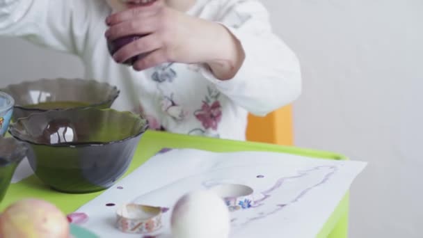 Beyaz gömlekli küçük bir kız tavuk yumurtalarını gıda boyasıyla boyuyor. Paskalya hazırlığı. Kapalı mekanda ellerle resim yapmak. 3 yaşından küçük çocuklar için yaratıcı bir gelişme. Eğitim, evde çizim yaparken — Stok video