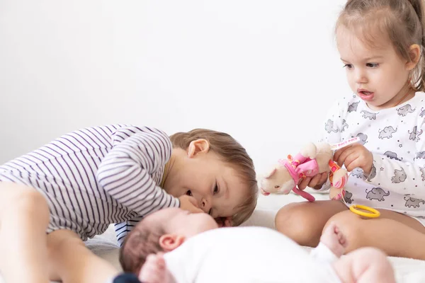 Piccoli bambini sorridenti che giocano insieme seduti sul letto. Fratello e sorella mostrano a un neonato un giocattolo. I bambini piccoli incontrano il fratellino appena nato. Bambino che dorme in buttafuori bianchi sotto una coperta. Bambini che giocano — Foto Stock