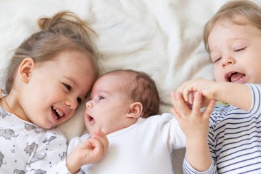 Yatakta birlikte oynayan gülümseyen küçük çocuklar. Erkek ve kız kardeş yeni doğmuş bir bebeğe oyuncak gösterir. Küçük çocuklar yeni doğan kardeşleriyle tanışıyor. Beyaz bir fedaide battaniyenin altında uyuyan bir bebek. Oyun oynayan çocuklar