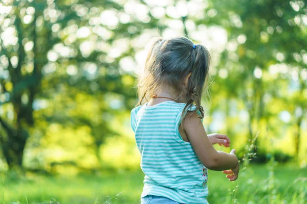 Jeugd, zomer en vrije tijd concept.Een schattige gelukkige kleine baby staat in helder gras met paardebloemen in de achtergrond van een zonsondergang in het park. meisje draaide haar rug besteden tijd buiten.Copy ruimte — Stockfoto