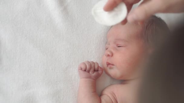 Infanzia, infanzia, maternità, igiene, medicina e concetto di salute - le mamme puliscono il viso del neonato nudo con un batuffolo di cotone sdraiato sul retro con l'ombelico bloccato da clip medica — Video Stock