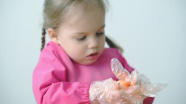 Pascua, Pascua, infancia, creatividad, concepto de juegos - primer plano de la pequeña rubia de tres años de edad, niña de aspecto caucásico esloveno en bata rosa pinta huevos de Pascua en guantes de plástico. — Vídeo de stock