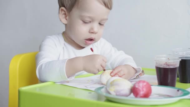 En liten blond pojke målar hönsägg genom att färga maten. Förberedelser inför påsk. Målning med händer inomhus. Kreativ utveckling för barn under 3 år. Utbildning, teckning hemma under — Stockvideo