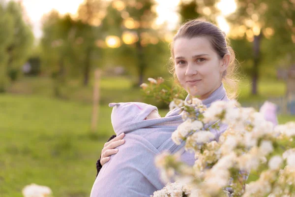 Maternidad, cuidado, bebés, verano, concepto de crianza - Joven hermosa mamá con un bebé recién nacido en una honda paseos y mira a un niño en la luz de fondo de una puesta de sol en el parque cerca del arbusto floreciente. — Foto de Stock