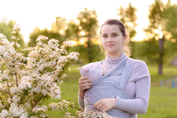 Maternità, cura, neonati, estate, concetto genitoriale - Giovane bella mamma con un neonato in fionda cammina e guarda un bambino nella retroilluminazione di un tramonto nel parco vicino al cespuglio fiorito. — Foto Stock