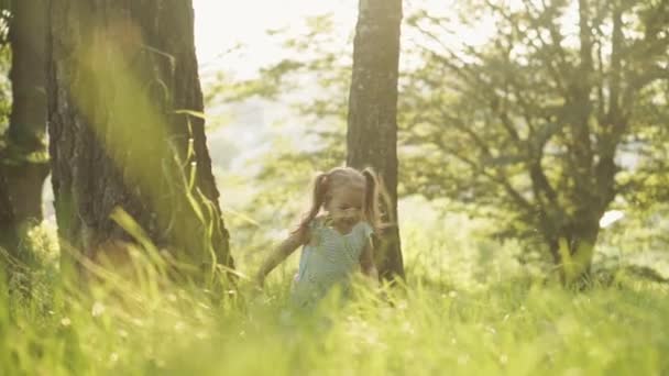 Ein kleines Mädchen rennt im Wald in die Kamera. Glücklich lächelnd versteckt sich das Kind bei Sonnenuntergang hinter den grünen Bäumen im Park. Sommer, Unterhaltung, Familie, Kindheit, Wandern, Urlaub, Lifestyle-Konzepte. — Stockvideo