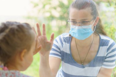 Maskeli Aile Anne ve Kızı. Virüse karşı koruyucu maskelerle pencerenin kenarında oturan mutlu genç bir kadın ve çocuk. Bebek cam üzerinde el ele tutuşuyor. Annem bebek yüzünü okşar.