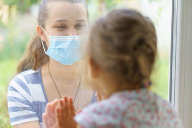 Maskeli Aile Anne ve Kızı. Virüse karşı koruyucu maskelerle pencerenin kenarında oturan mutlu genç bir kadın ve çocuk. Bebek cam üzerinde el ele tutuşuyor. Annem bebek yüzünü okşar.