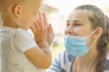 Maskeli Aile Anne ve Kızı. Virüse karşı koruyucu maskelerle pencerenin kenarında oturan mutlu genç bir kadın ve çocuk. Bebek cam üzerinde el ele tutuşuyor. Annem bebek yüzünü okşar.
