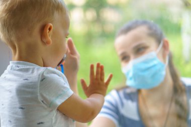 Maskeli Aile Anne ve Kızı. Virüse karşı koruyucu maskelerle pencerenin kenarında oturan mutlu genç bir kadın ve çocuk. Bebek cam üzerinde el ele tutuşuyor. Annem bebek yüzünü okşar.