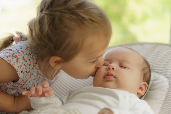 Милая маленькая девочка целует его новорожденную сестру. Малыш Тоддлер знакомится с новорожденным братом. Младенец спит в белом вышибале под одеялом. Дети играют и общаются. Дети с небольшой разницей в возрасте. — стоковое фото