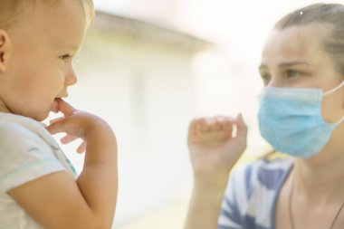 Maskeli Aile Anne ve Kızı. Virüse karşı koruyucu maskelerle pencerenin kenarında oturan mutlu genç bir kadın ve çocuk. Anne ve bebek cam üzerinde el ele tutuşup birbirlerine bakıyorlar.