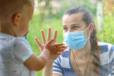 Maskeli Aile Anne ve Kızı. Virüse karşı koruyucu maskelerle pencerenin kenarında oturan mutlu genç bir kadın ve çocuk. Anne ve bebek cam üzerinde el ele tutuşup birbirlerine bakıyorlar.
