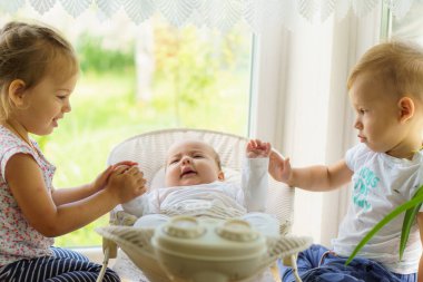 Pencerenin yanında üç küçük çocuk oynuyor. Bebekli kız ve erkek kardeşler. Bebek yeni doğan kardeşiyle tanışıyor. Küçük yaş farkı olan çocuklar el ele tutuşur, öpüşür, daha genç ailelerle eğlenir.