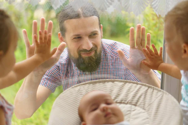 Famiglia è divisa in epidemia. giovane padre guarda i minori attraverso la finestra tiene la mano sul vetro. simbling con piccola differenza di età.Due bambini con neonato seduto vicino alla finestra sulla — Foto Stock