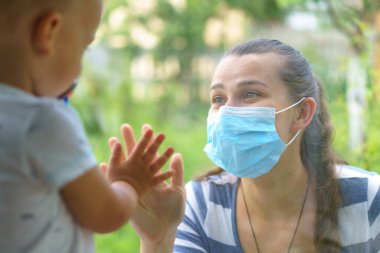 Tıbbi Maskeli Anne ve Kızı Ailesi. Virüse karşı koruyucu maskeli pencerenin yanında oturan genç kadın ve çocuk. Anne ve bebek cam üzerinde el ele tutuşup birbirlerine bakıyorlar. Yüksek kalite fotoğraf