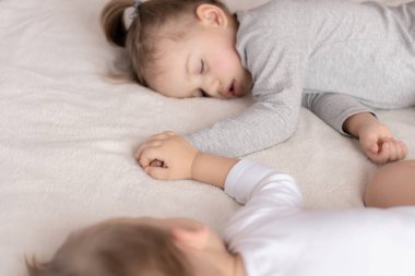 Çocukluk, uyku, rahatlama, aile, yaşam tarzı konsepti. 2 ve 3 yaşlarındaki iki küçük çocuk beyaz ve bej renkli giysiler içinde öğle yemeğinde el ele tutuşarak bej ve beyaz bir yatakta uyurlar.