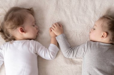 Çocukluk, uyku, rahatlama, aile, yaşam tarzı konsepti. 2 ve 3 yaşlarındaki iki küçük çocuk beyaz ve bej renkli giysiler içinde öğle yemeğinde el ele tutuşarak bej ve beyaz bir yatakta uyurlar.