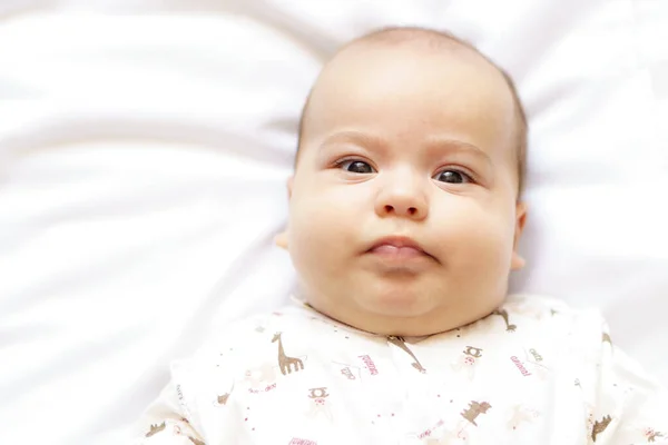 Μικρό παχουλό νεογέννητο μωρό με λευκή φόρμα ξαπλωμένο στο κρεβάτι. Κάτοψη ενός λυπημένου νεογέννητου μωρού σε μια λευκή ζεστή κουβέρτα. Η φοβισμένη απογοητευμένη υπνηλία νεογέννητο με καστανά μάτια είναι ξύπνιος κοιτάζοντας γύρω — Φωτογραφία Αρχείου