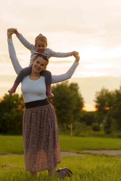 Moederschap, zorg, baby 's, zomer, ouderschap concept - Jonge moeder met drie jaar oud dochtertje in haar armen loopt en kijkt naar het kind in de achtergrond van zonsondergang in het park in de buurt van bloeiende struik. — Stockfoto