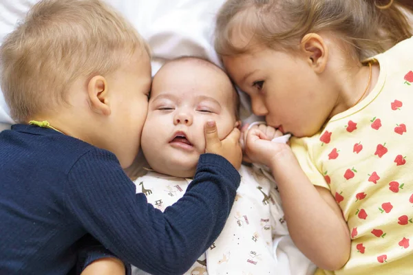 Kleine lächelnde Kinder, die zusammen auf dem Bett spielen. Bruder und Schwester küssen ein Neugeborenes. Kleinkind trifft neugeborenes Geschwisterchen. Säugling schläft in weißem Türsteher unter einer Decke. Kinder mit kleinem Alter — Stockfoto