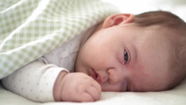 Kojenectví, dětství, vývoj, medicína a zdraví koncept - close-up tvář novorozence baculaté spící dítě dívka ležící na břiše otevřené oči a úsměv pokrytý přikrývkou na posteli na bílém pozadí. — Stock video