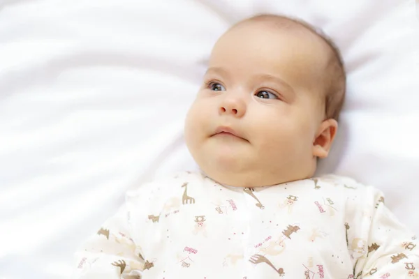 Little newborn baby in a white bodysuit lying on the bed. Top view of a newborn baby smiles on a white warm blanket. The newborn is awake looking around indoors