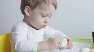 Sarışın bir çocuk tavuk yumurtalarını gıda boyasıyla boyuyor. Paskalya hazırlığı. Kapalı mekanda ellerle resim yapmak. 3 yaşından küçük çocuklar için yaratıcı bir gelişme. Eğitim, evde çizim yaparken