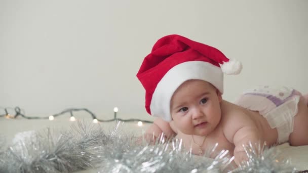 Wesołych Świąt Bożego Narodzenia i szczęśliwego Nowego Roku, niemowlęta, dzieciństwo, koncepcja świąt - zbliżenie nagie 6 miesięcy noworodka w kapeluszu Świętego Mikołaja na brzuchu roi się od dekoracji na choince. — Wideo stockowe