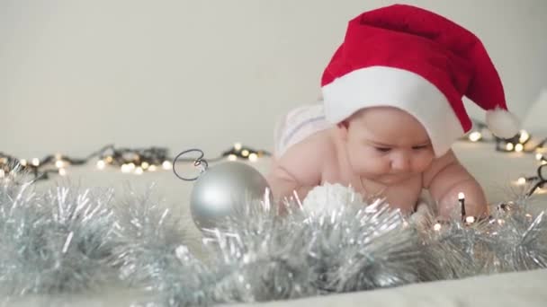 Merry christmas christmas and happy new year, infants, childhood, holidays concept - close-up naked 6 month old newborn baby in santa claus hat on his tummy crawls with decorations on christmas tree. — Stock Video