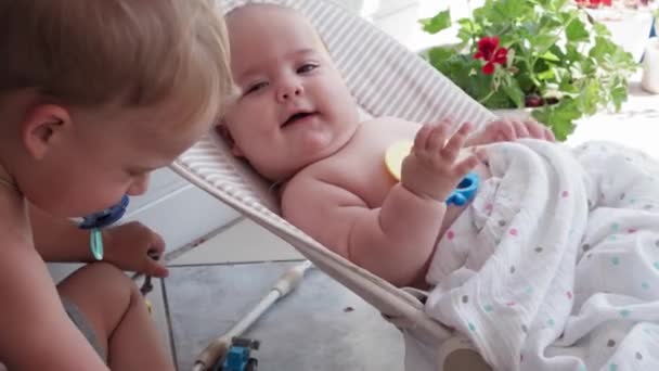 Βρεφική ηλικία, παιδική ηλικία, ανάπτυξη, καλοκαιρινές διακοπές, ιατρική και υγεία έννοια - Close-up top view πορτρέτο του νεογέννητου κοριτσιού πέντε μηνών που βρίσκεται σε κουνιστή καρέκλα Μεγαλύτερος αδελφός δώσει παιχνίδι έξω — Αρχείο Βίντεο