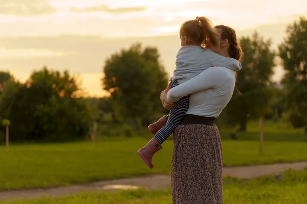 Maternità, cura, neonati, estate, concetto di genitorialità - Giovane mamma con tre anni bambina figlia tra le braccia cammina e guarda il bambino in controluce del tramonto nel parco vicino cespuglio fiorito. — Foto Stock