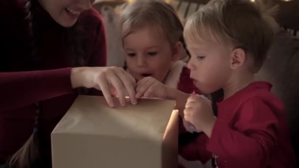 Xmas, vinter, nyår, fest, familj koncept - Ung lycklig mamma med två små barn pojke och flicka klädd i röda kläder öppna presentförpackning sitta nära dekorerad julgran. starkt ljus från den — Stockvideo