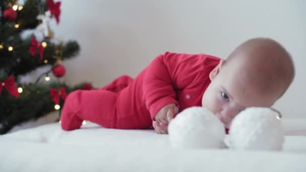 Весёлого Рождества и с Новым годом, младенцы, детство, праздничная концепция - крупным планом 6-месячный новорожденный младенец в шляпе Санта-Клауса и красном купальнике на животе ползает с шарами украшения на рождественской елке — стоковое видео