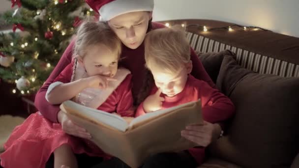 Xmas, vinter, nytt år, fest, familj koncept - Ung lycklig mamma två små barn pojke och flicka klädd i röda kläder läsa sagor sitta nära dekorerad julgran starkt ljus från boken — Stockvideo