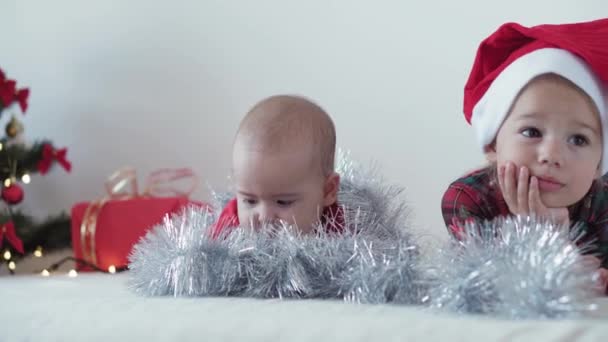 Feliz Natal e feliz ano novo, criança, infância, conceito de férias - close-up bebê recém-nascido de 6 meses em chapéu de santa claus e bodysuit vermelho com a irmã mais velha rasteja com decorações árvore de Natal bola — Vídeo de Stock